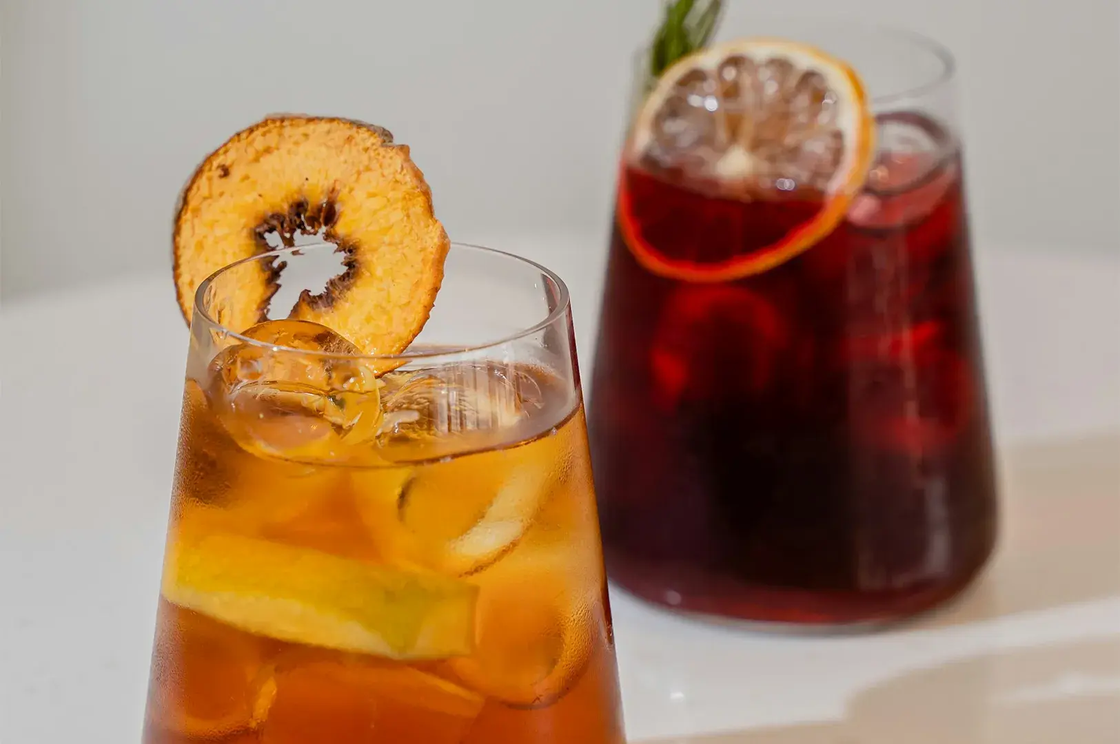 Two glasses of iced beverages against a light background: one amber drink garnished with a dried fruit slice, and a deeper red drink garnished with a dried citrus wheel.