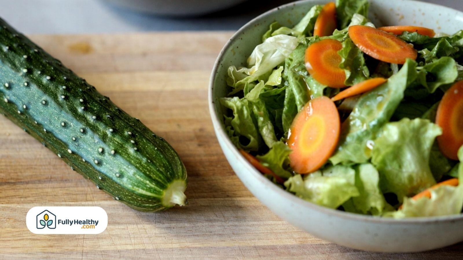 Fresh whole cucumber next to a bowl of lettuce and sliced carrots.