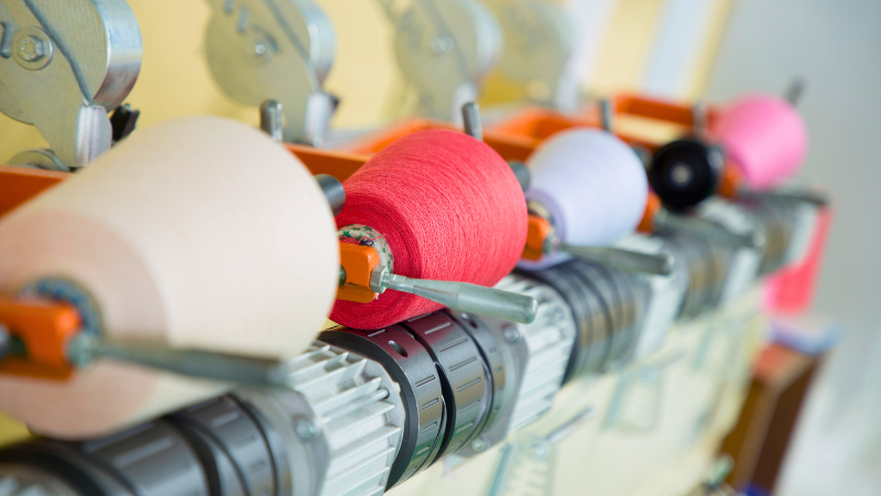 Spools of recycled polyester thread in colors like cream, red, white, black, and pink on a textile machine.