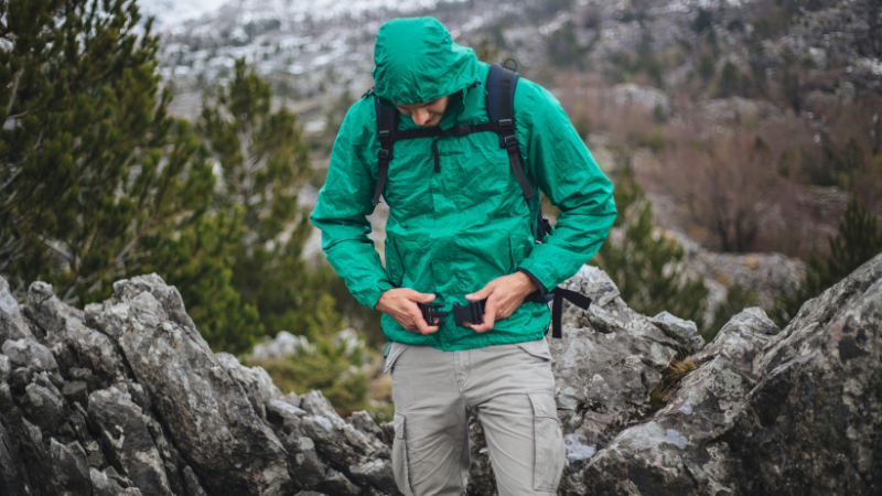 A person in a green gorpcore rain jacket adjusts a backpack strap on rocky ground.