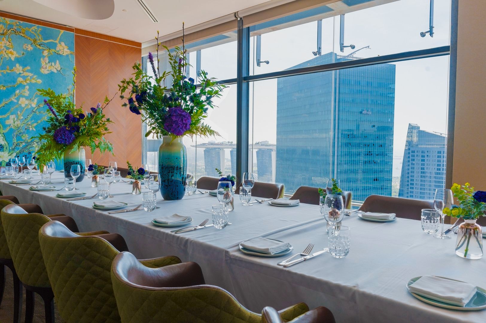 Elegant dining room with a long table set for a banquet. Large blue vases with purple flowers are placed along the table. Floor-to-ceiling windows offer a city skyline view.