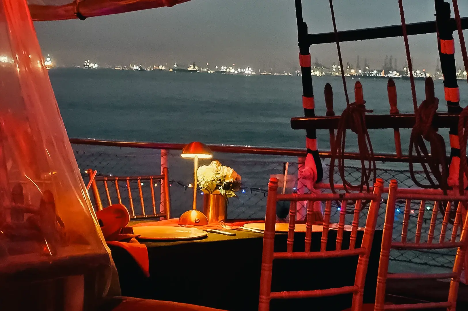 Eye‑level evening shot of an outdoor fine‑dining table set on a boat or waterfront deck, with candlelight, fabric chairs, and city lights reflected across calm water.
