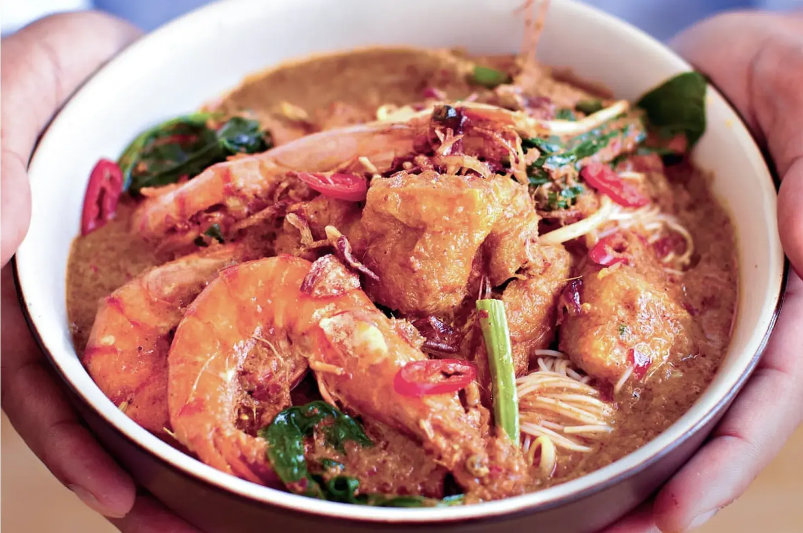 A close-up of a bowl of seafood laksa, held in hands. The dish features vibrant prawns, noodles, and leafy greens in a rich, spicy broth.