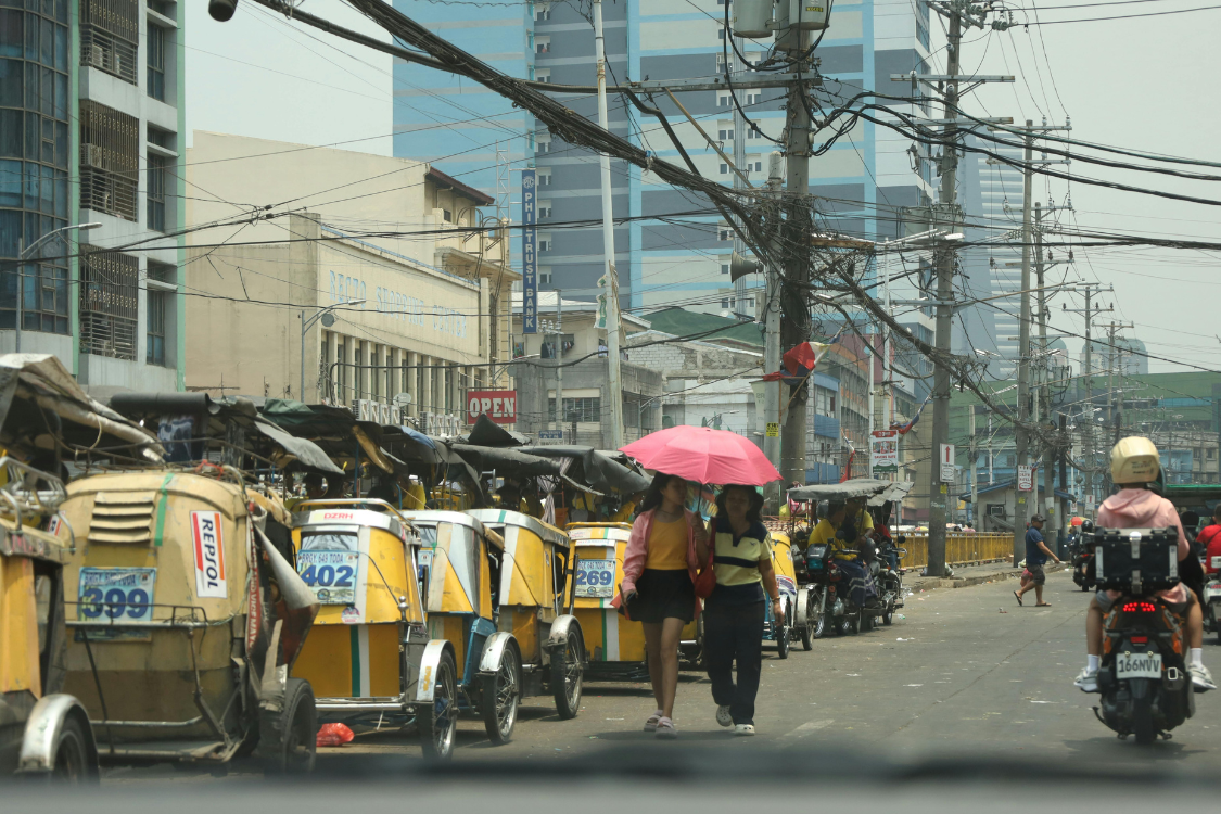 Street scene in the Philippines showing tricycles lined up, pedestrians with umbrellas, and motorcycles passing through a busy urban area.
