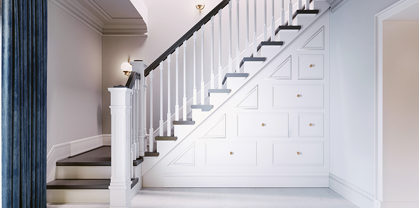 This image displays a mostly-white staircase against beige walls and an ocean-blue floor-to-ceiling curtains. The side of the staircase features multiple storage cupboards, the dark wooden steps and handrail contrasted with the white balustrades. Gold wall sconces and detailed wall trimmings feature to bring different textures to the room.