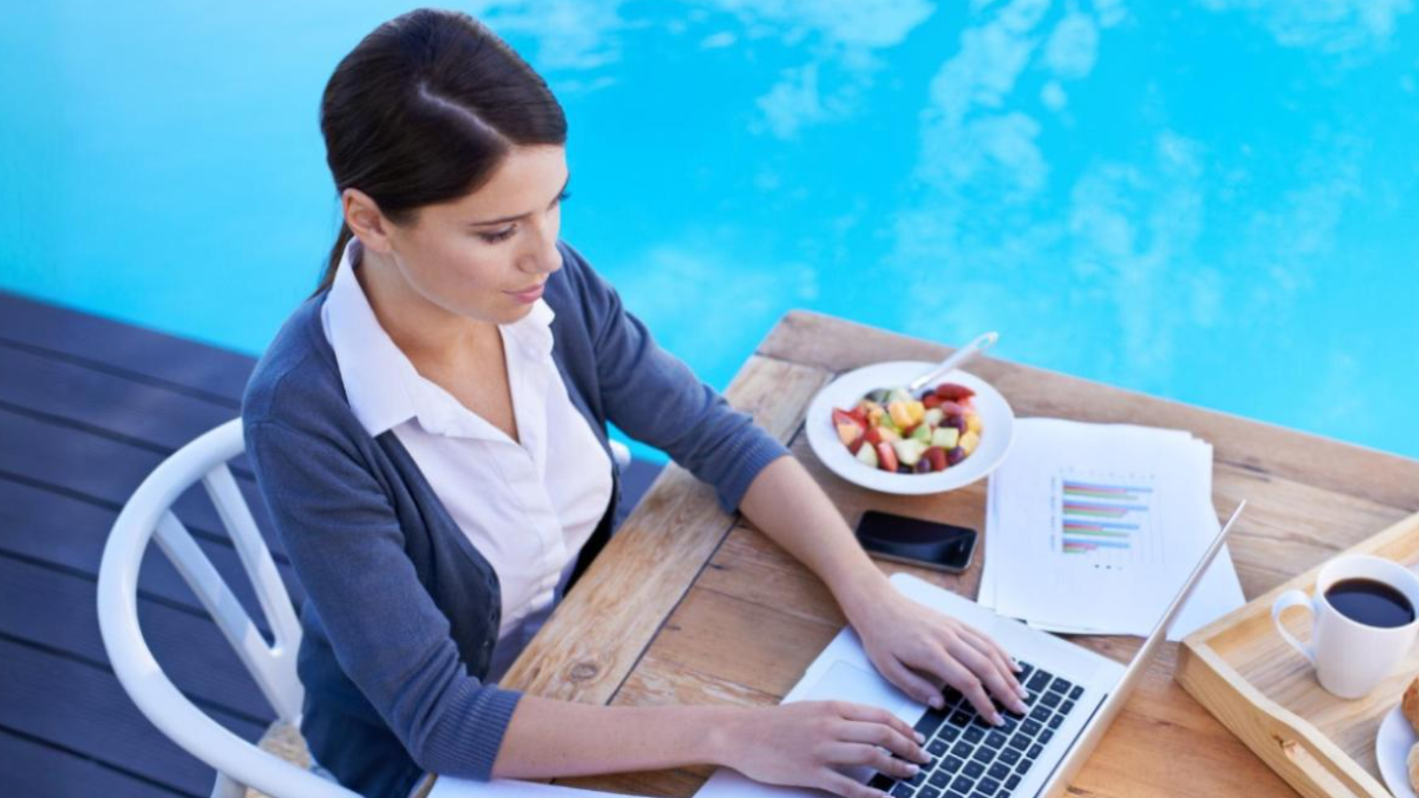 Person working on a laptop beside a pool with charts, coffee, and a fruit bowl nearby, illustrating remote working with cloud PBX (Private Branch Exchange)