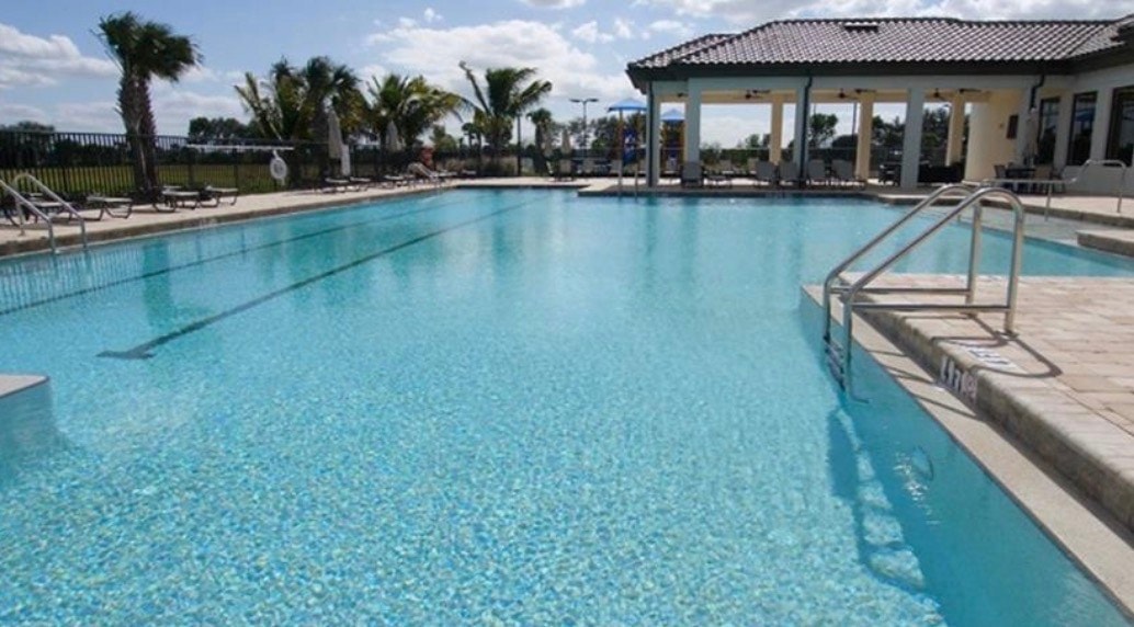The image depicts the resort-style pool area at Portico, featuring lap lanes and a spacious sun deck, with a modern clubhouse in the background surrounded by lush palm trees. This inviting space highlights the exquisite modern luxury and amenities available in the highly desired gated community of Portico in Fort Myers, FL.