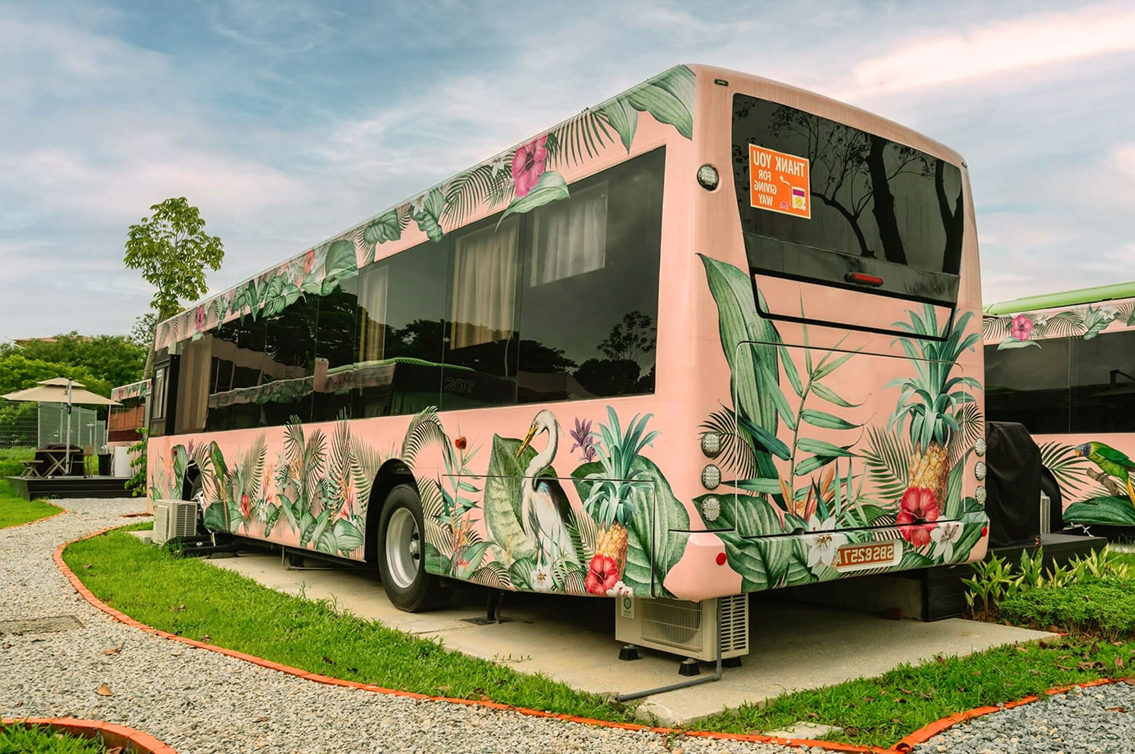 A stationary pink bus with tropical leaves and toucan artwork. It sits by a gravel path under a cloudy sky, creating a cheerful, whimsical scene.