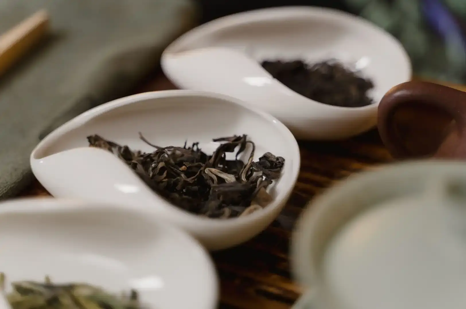 A soft-focus, intimate detail shot of a white tea-serving tray, featuring teardrop-shaped dishes containing various loose-leaf teas, with the warm, rich tones of the tea leaves contrasting against the clean white ceramic.
