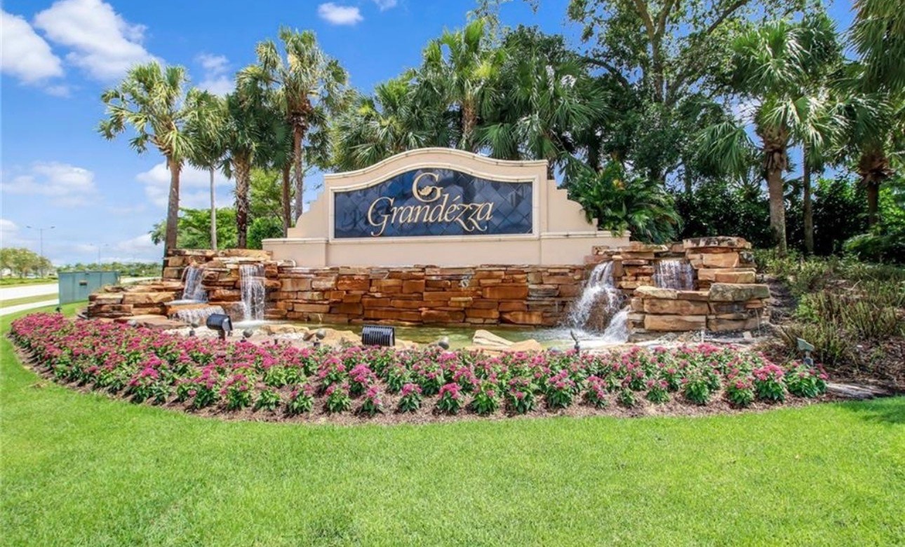 The image depicts a luxurious single-family home located in a gated community in Estero, FL, featuring stunning views of the nearby golf course and meticulously maintained mature landscaping. This elegant residence is part of the Grandezza community, known for its beautiful homes and convenient access to amenities like tennis courts and a clubhouse.
