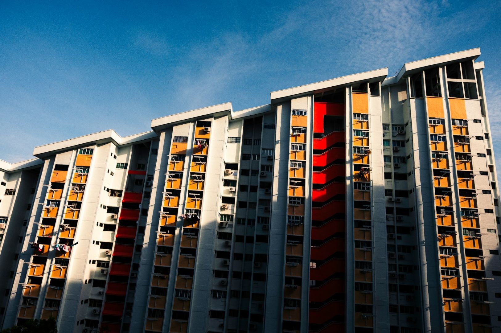 Sunlit high-rise building with white, orange, and red panels under a clear blue sky. Shadows cast diagonally create contrast and depth.