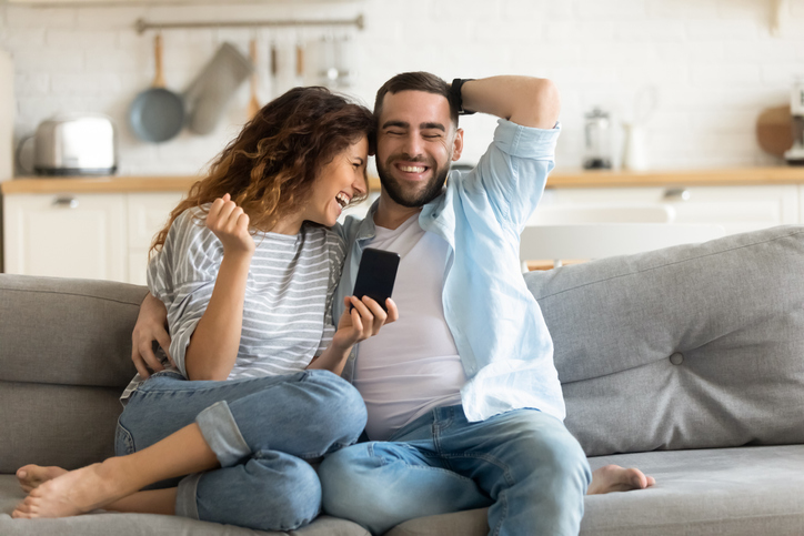 Happy young couple sitting on a gray sofa looking at a cell phone.  