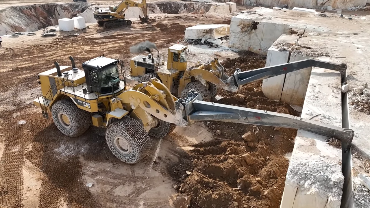 Two large yellow Caterpillar wheel loaders working in a quarry, one is transporting dirt while the other lifts a heavy block of stone using a metal arm attachment