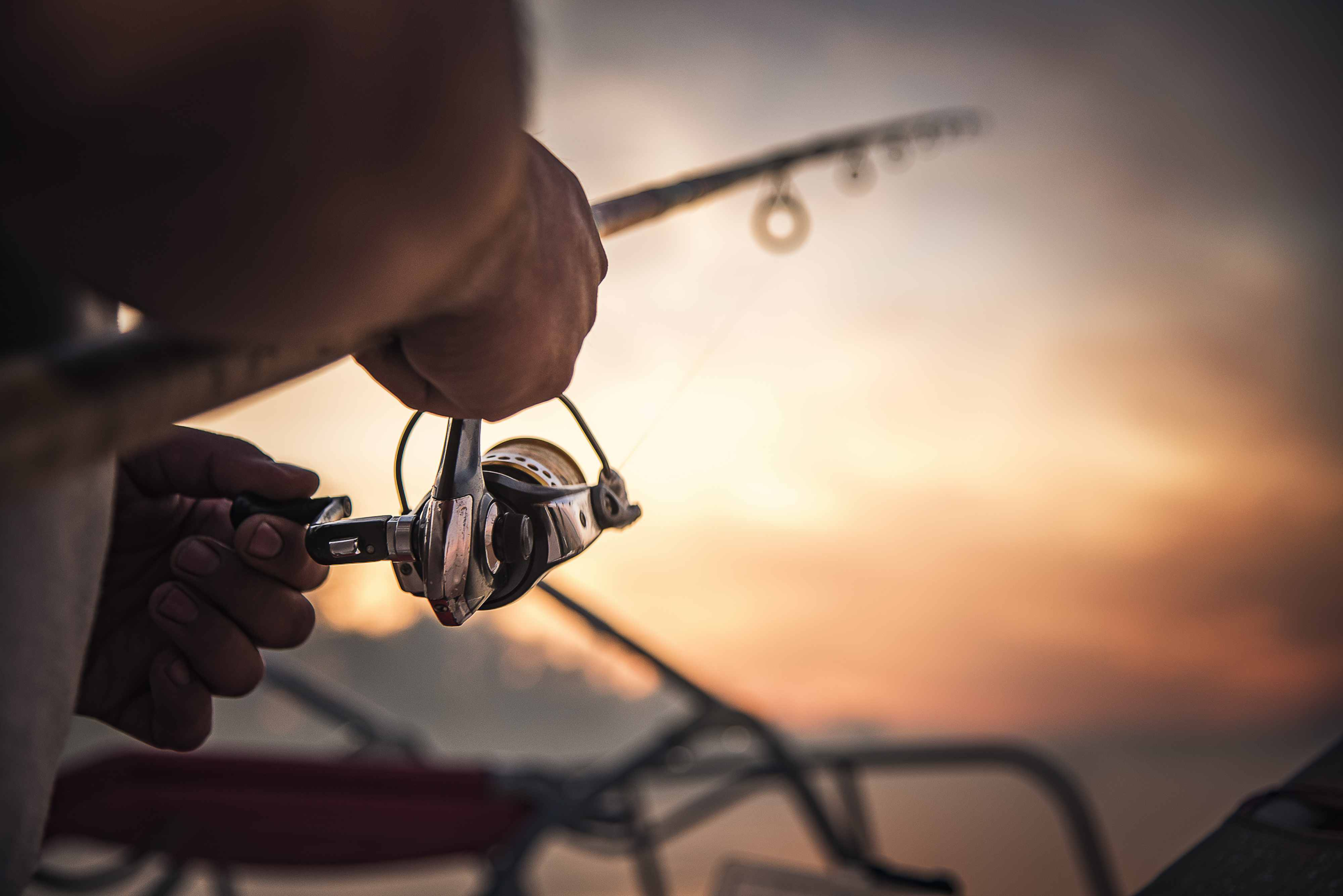 Carretel de pesca em close-up, pescador segurando vara de pesca, fundo pôr do sol