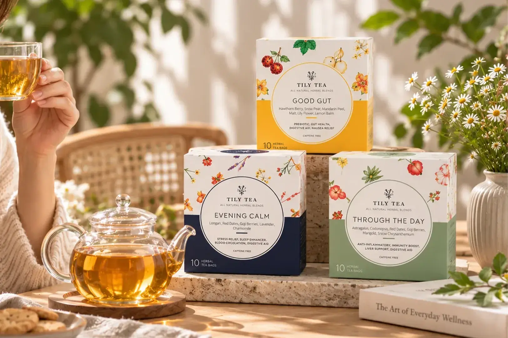 Lifestyle product setting featuring three Tily Tea herbal tea boxes, a glass teapot filled with amber tea, and a hand holding a glass cup.