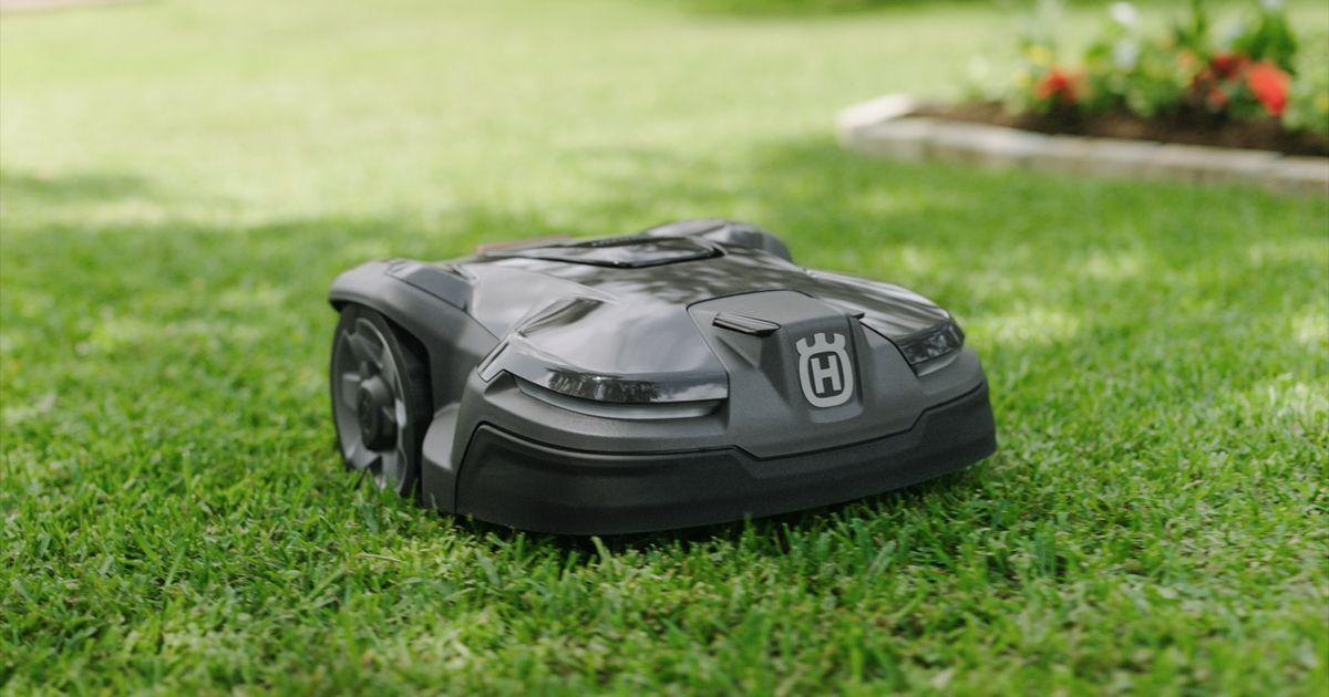 A black Husqvarna robotic lawn mower is autonomously cutting grass on a well-manicured lawn. The mower has a sleek design and operates near a flower bed with red and yellow flowers. The background is slightly blurred, focusing on the mower's activity.
