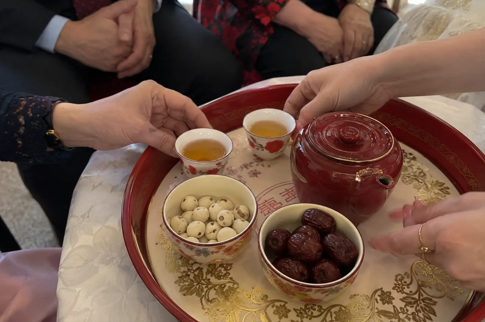 A close-up shot focuses on a round red lacquer tray adorned with gold floral patterns and a faint "Double Happiness" Chinese character symbol in the center. Resting on the tray is a smooth, dark red teapot next to two small white ceramic teacups filled with amber-colored tea, each detailed with red flower motifs. Two small patterned bowls sit in the foreground, one filled with white candied lotus seeds and the other with wrinkled dark red jujubes. Multiple hands extend into the frame, with one hand gently holding a teacup and another resting on the edge of the tray against a background of wedding attire.