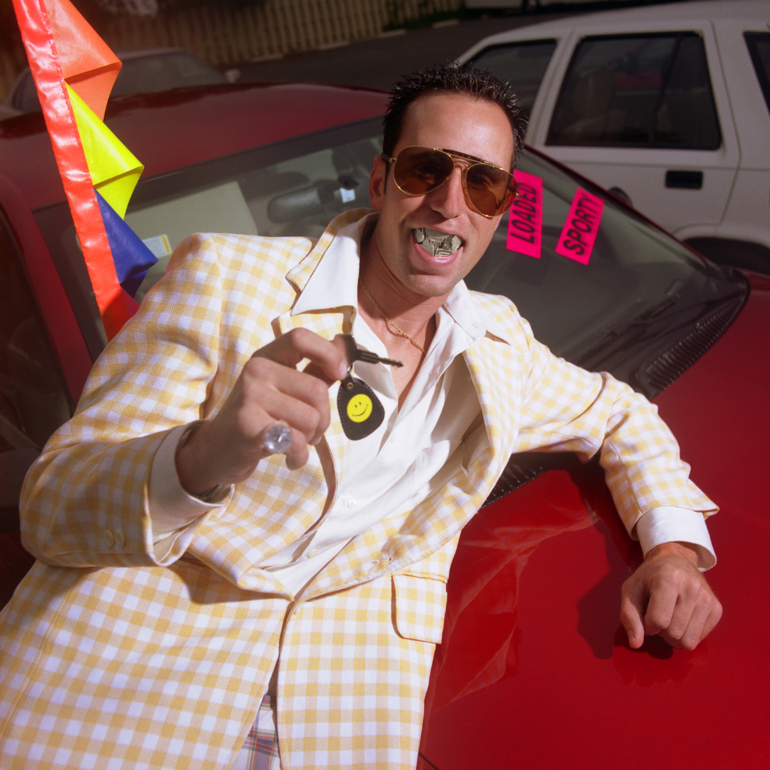 A guy in a cheap suit with "grill" style teeth leans on a car in a used car lot, trying to sell it to the gullible.