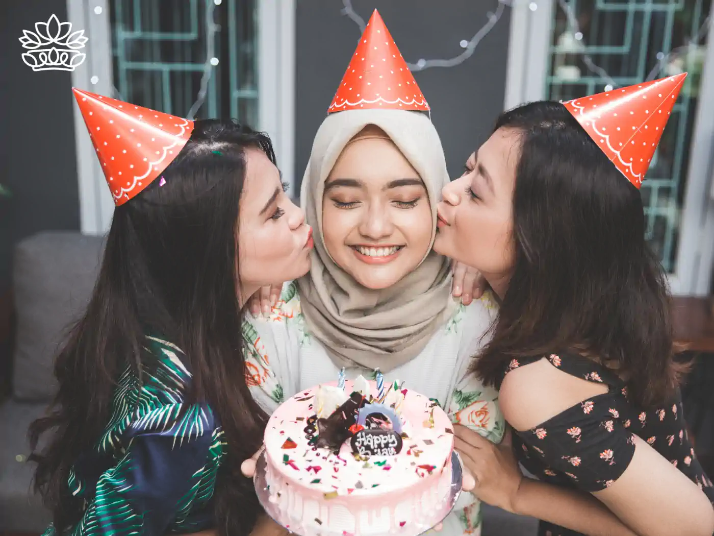 Two women wearing party hats kissing another woman on the cheeks as she holds a birthday cake, all smiling. Fabulous Flowers and Gifts.