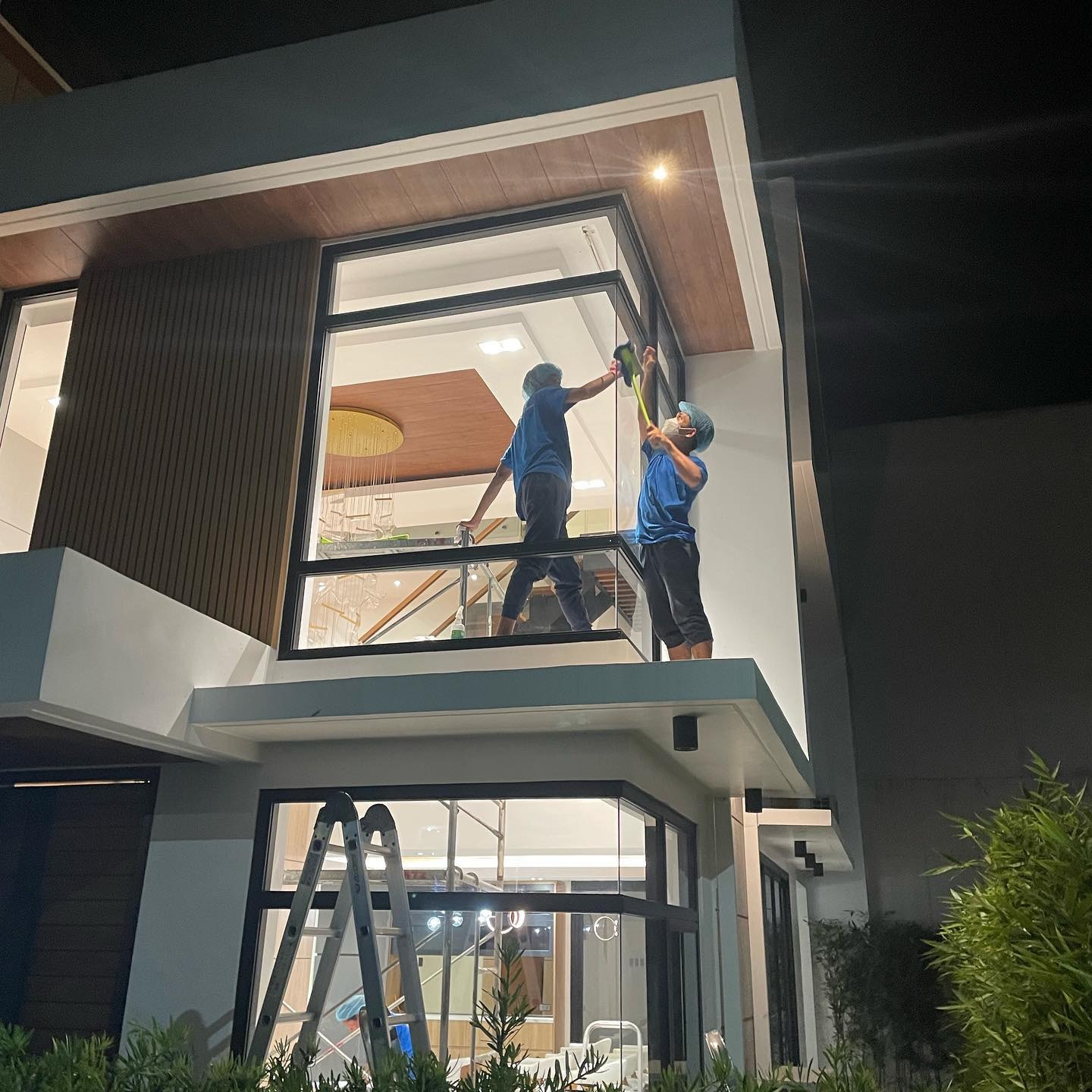 Two individuals in blue uniforms and protective gear cleaning large upper-level glass windows of a modern house at night. One person is standing on a balcony while the other uses a long-handled tool to reach the top edges.