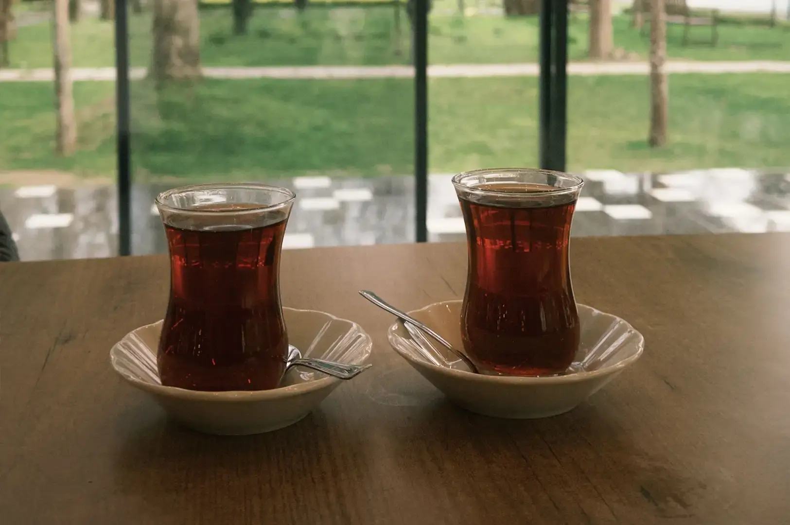 Two filled tulip-shaped glasses of hot Turkish tea, each accompanied by a tiny spoon and resting on off-white fluted saucers, sit side-by-side on a wooden table in front of a large window overlooking a lush green park scene.