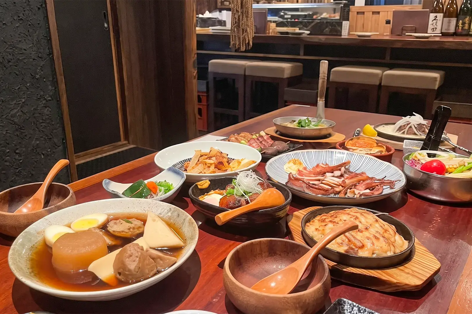 A table filled with a variety of Japanese izakaya dishes including oden, grilled squid, and hot plates in a restaurant setting.