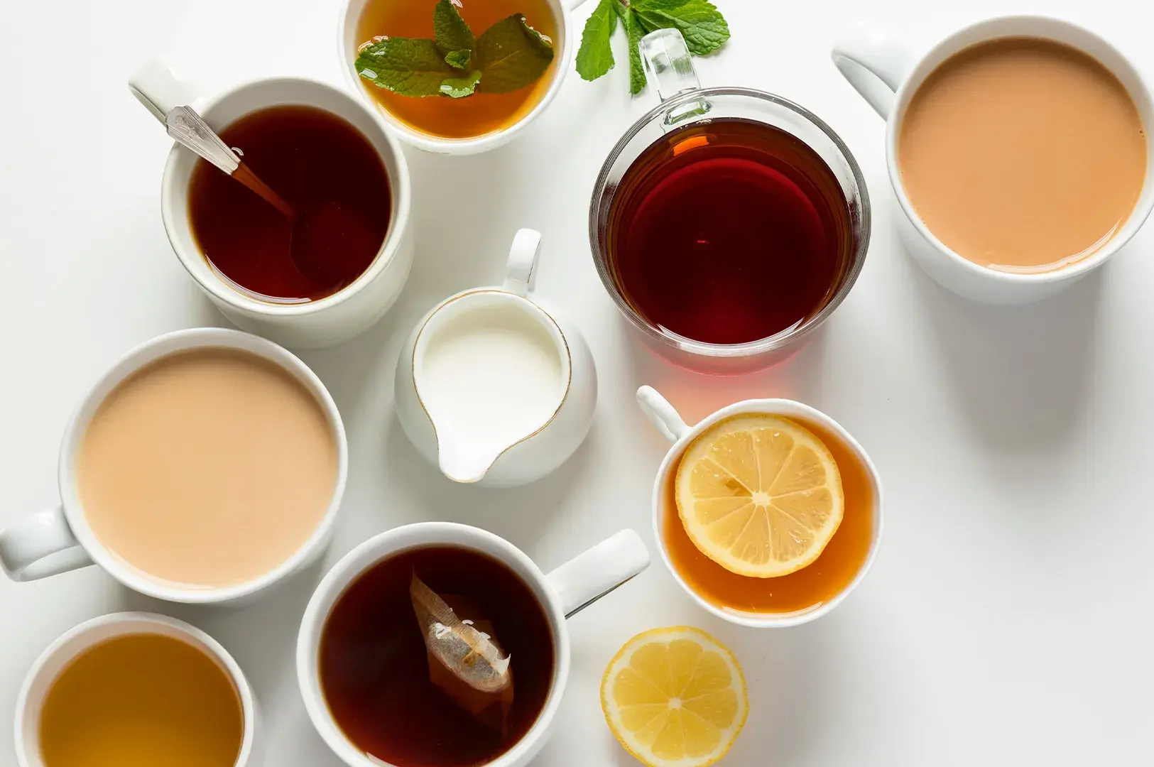 Flat lay of various cups of tea including milk tea, black tea, herbal tea with mint, and lemon tea arranged around a small milk pitcher on a white surface.