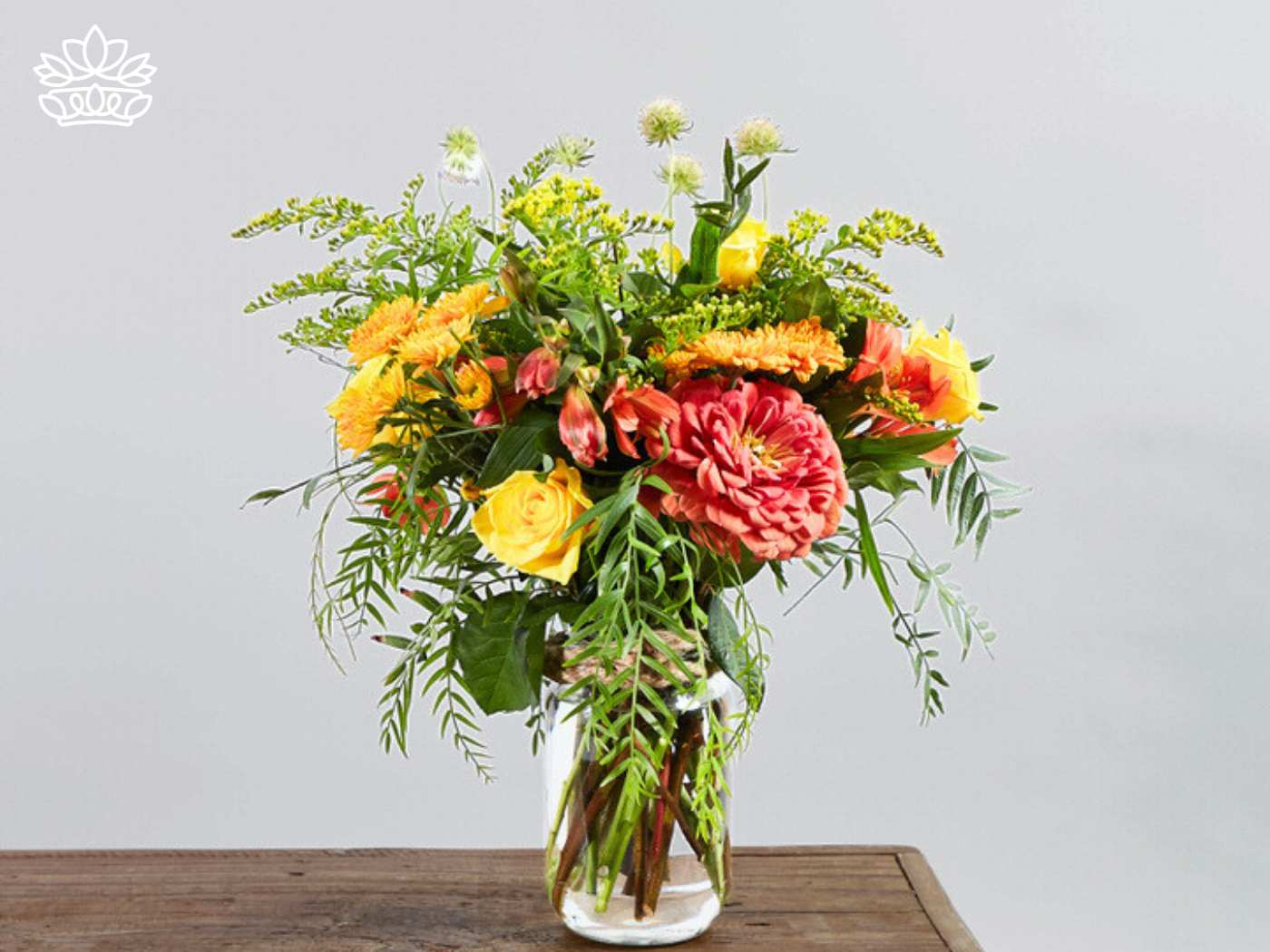 A vibrant floral arrangement of orange and yellow flowers with lush greenery, displayed in a clear glass vase on a wooden surface. Fabulous Flowers and Gifts. Gifts for Him. Delivered with Heart.