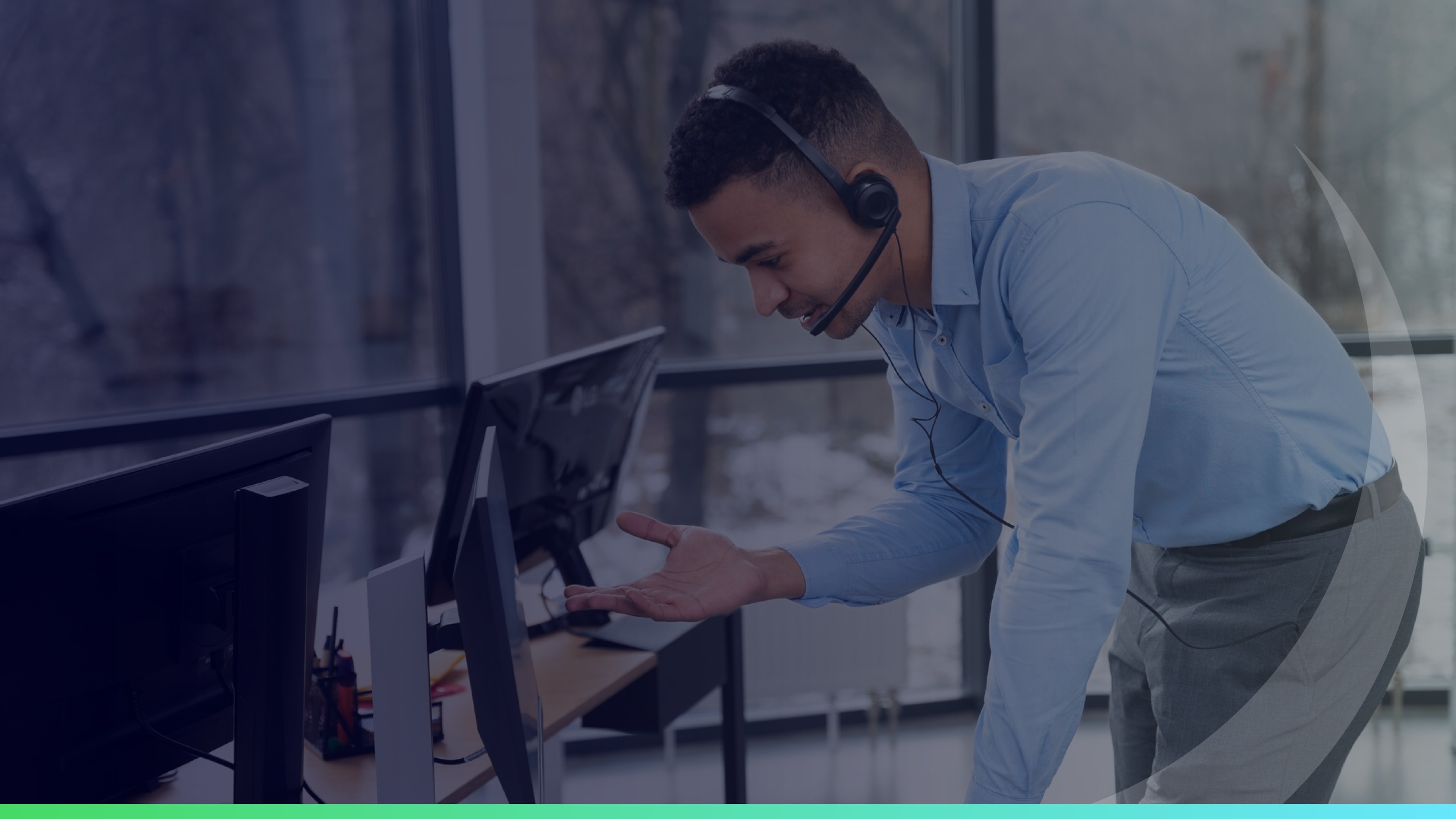 Man wearing a headset and gesturing while speaking to someone on a video call in an office with multiple monitors.