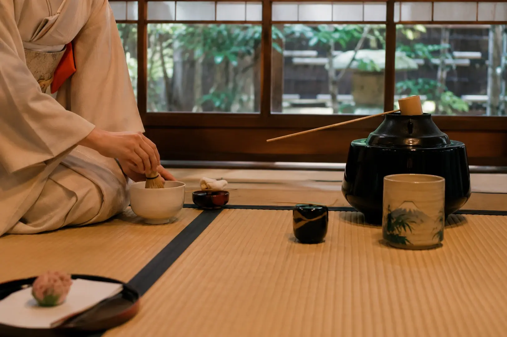 Close-up on the host's hands as she whisks tea in a white ceramic bowl, focusing closely on the ritual of the preparation. To the right stands a large black iron kettle with a bamboo ladle resting on top, while in the immediate left foreground sits a single pink wagashi sweet on a white paper. The background features open shoji doors looking out into a garden, grounding the intimate act of tea preparation in a larger, natural setting.
