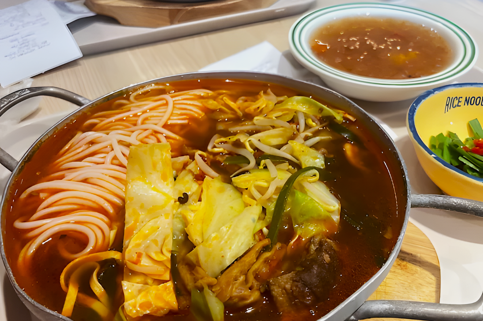 A steaming pot of spicy noodle soup filled with noodles, cabbage, bean sprouts, and meat sits on a table next to a bowl of sauce and garnishes.
