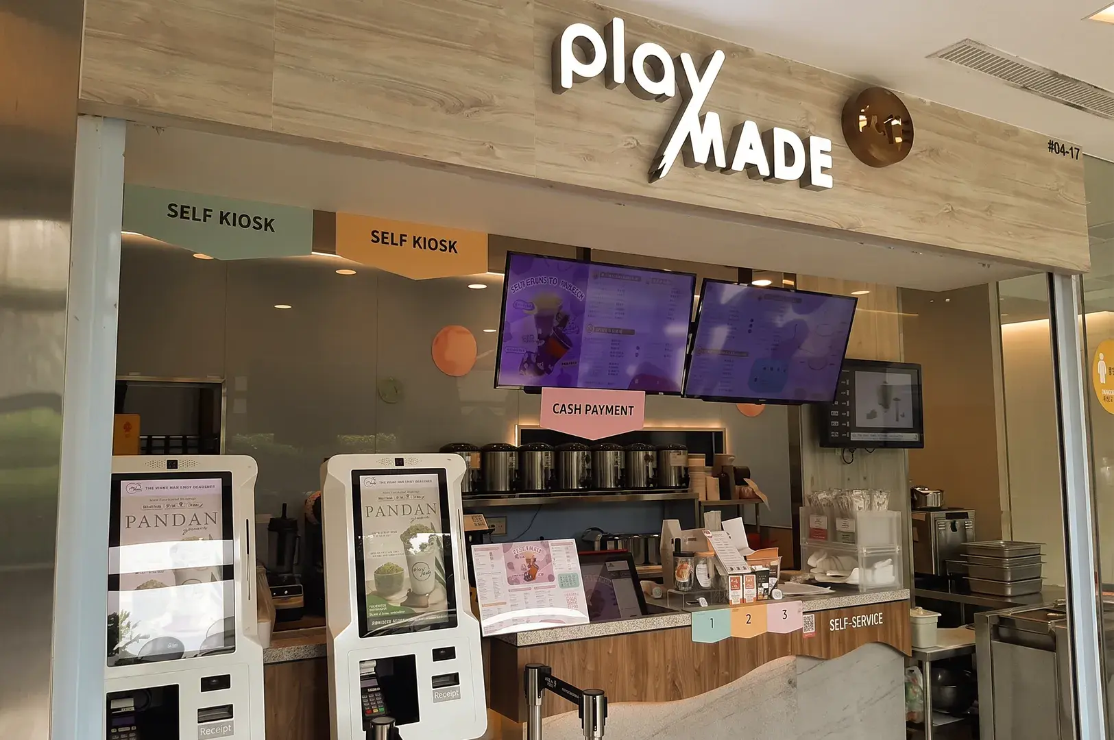 Storefront of a PlayMade beverage shop featuring self-service kiosks, digital menu screens, and a service counter.