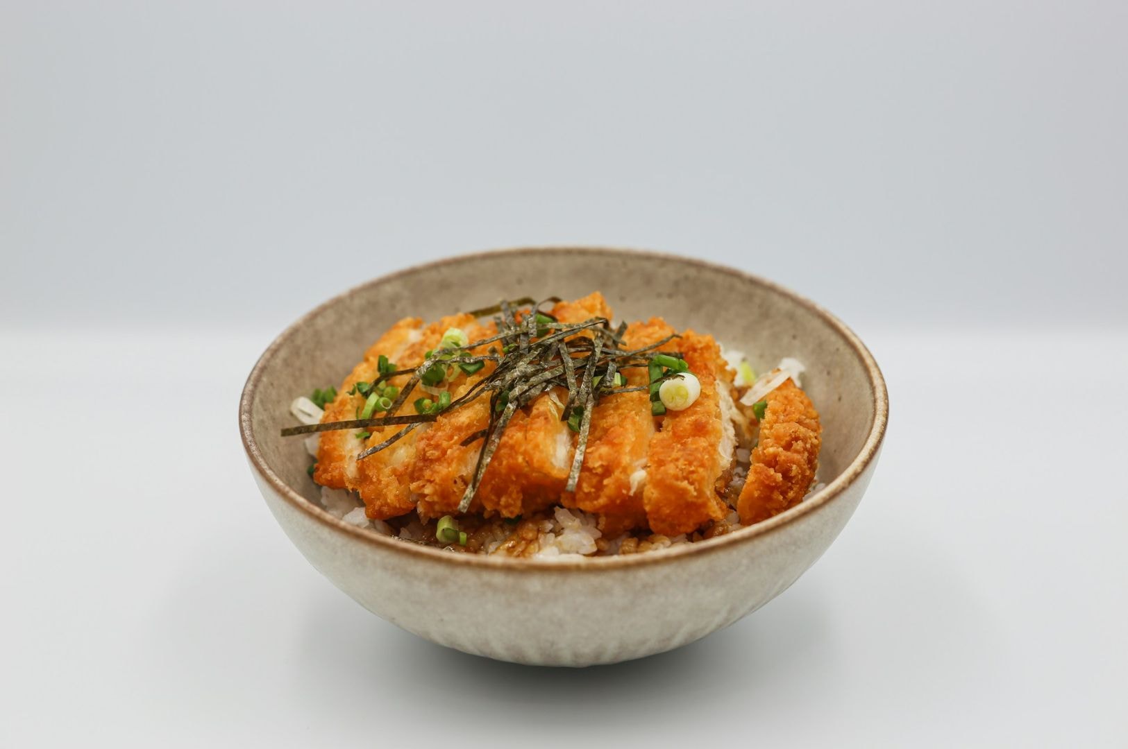 A bowl of crispy, golden chicken katsu slices on rice, topped with sliced green onions and seaweed strips. The dish has a fresh and appetizing appearance.