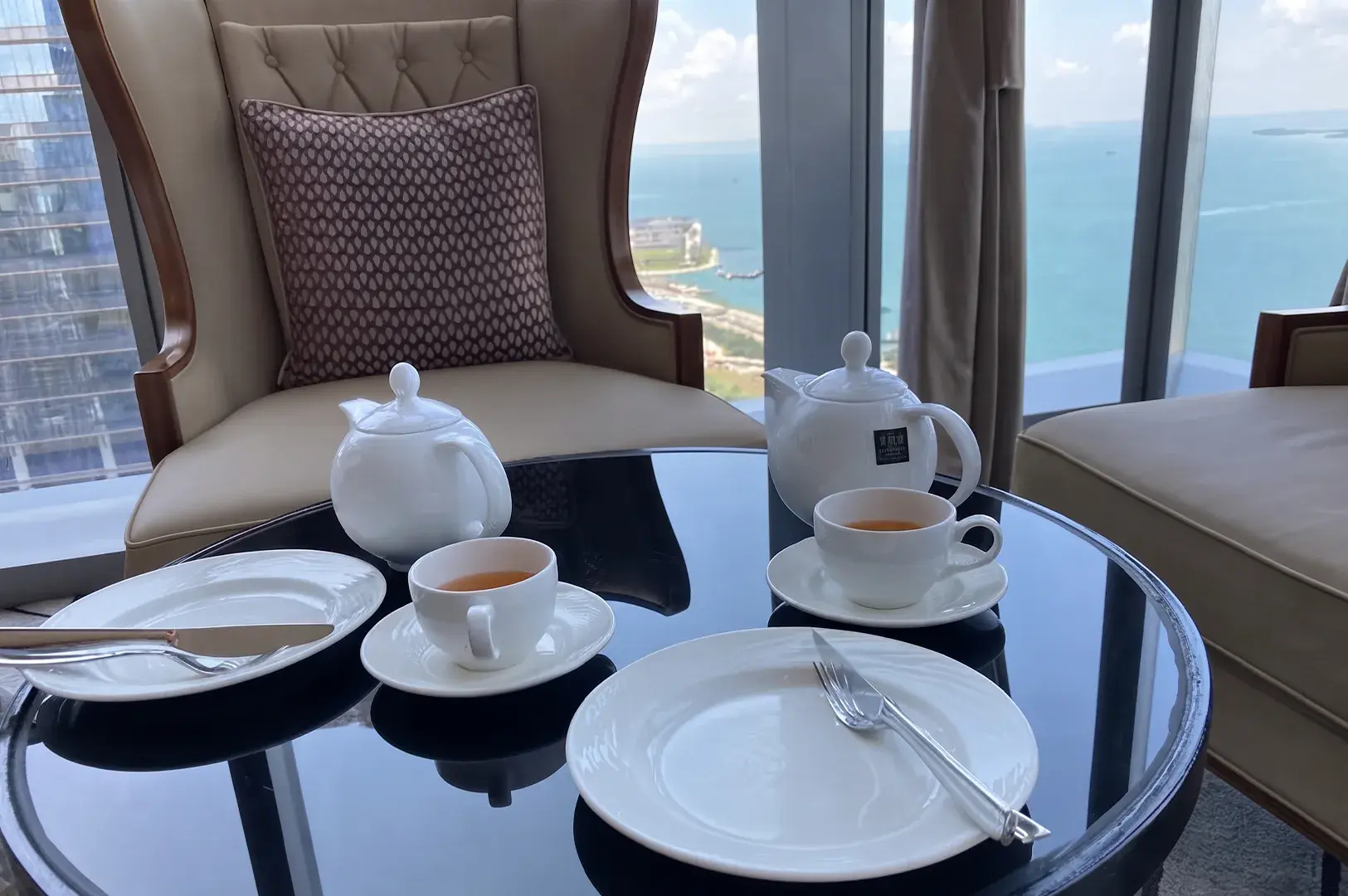 High tea table arrangement with white ceramic teapots, teacups, and empty plates set beside plush armchair seating overlooking a high-rise ocean view.