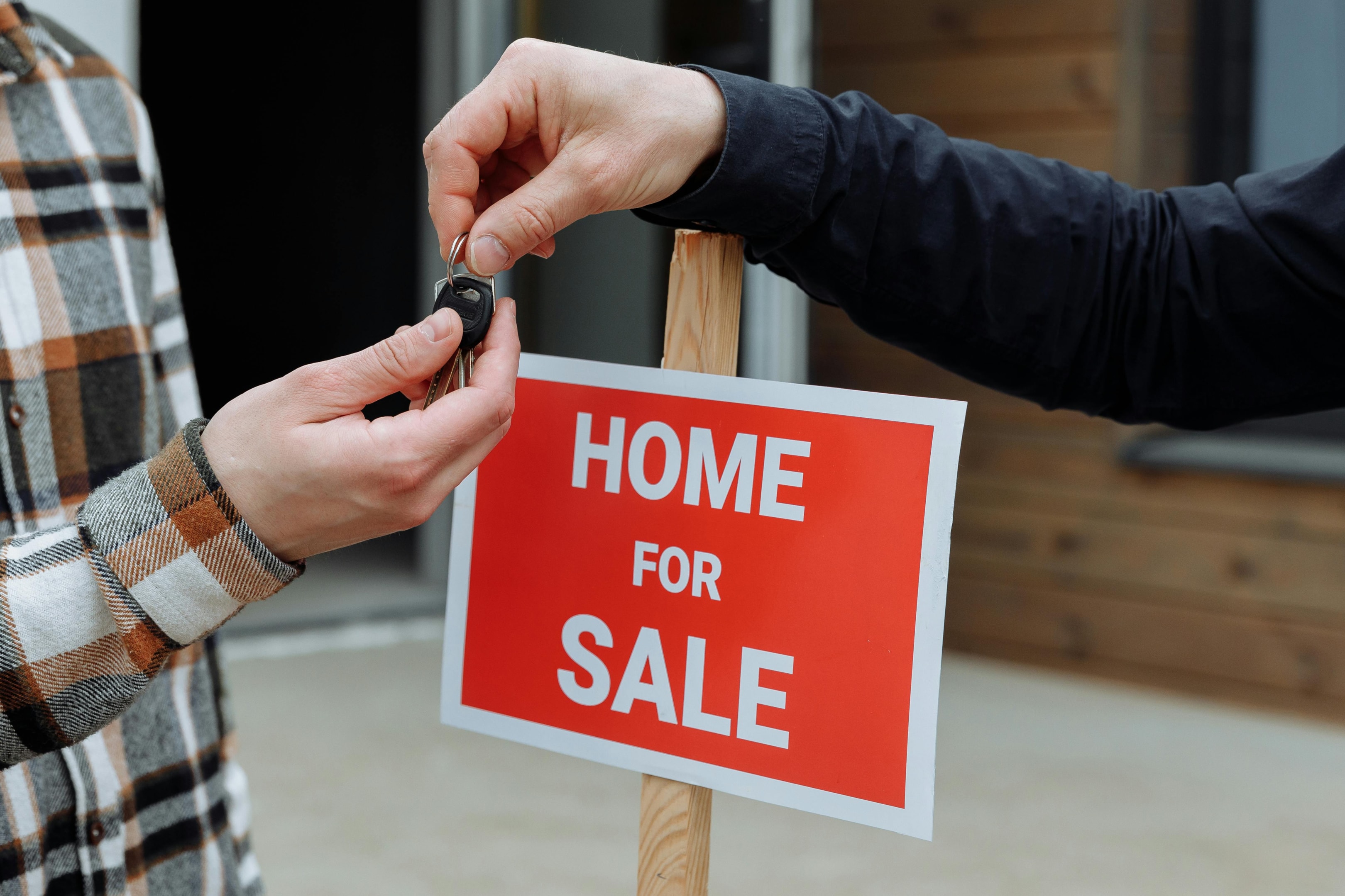 Keys being passed from real estate agent to home buyer in front of 'home for sale' sign.