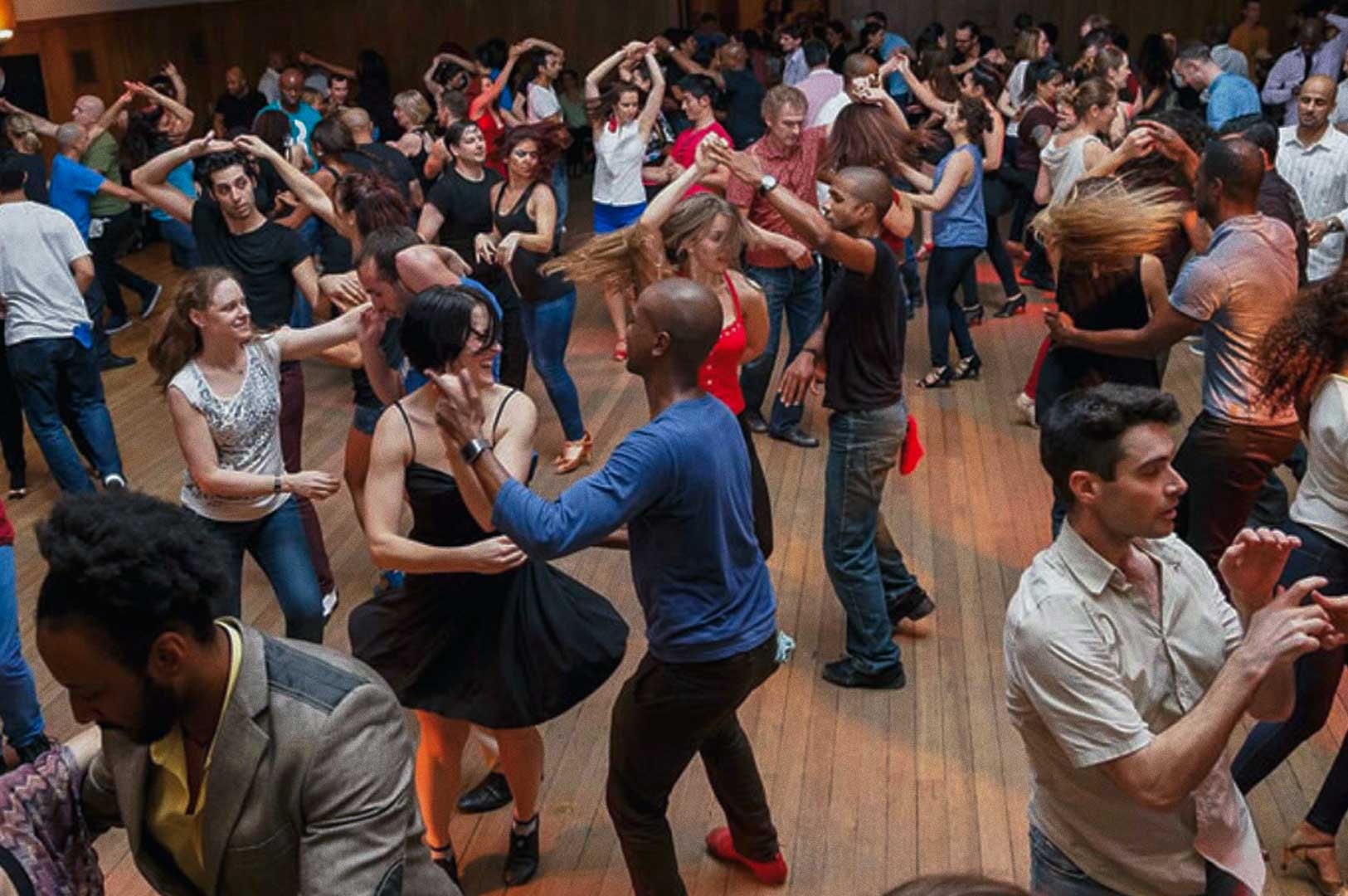 Crowd dancing at a lively social dance event in a large wooden-floor hall.