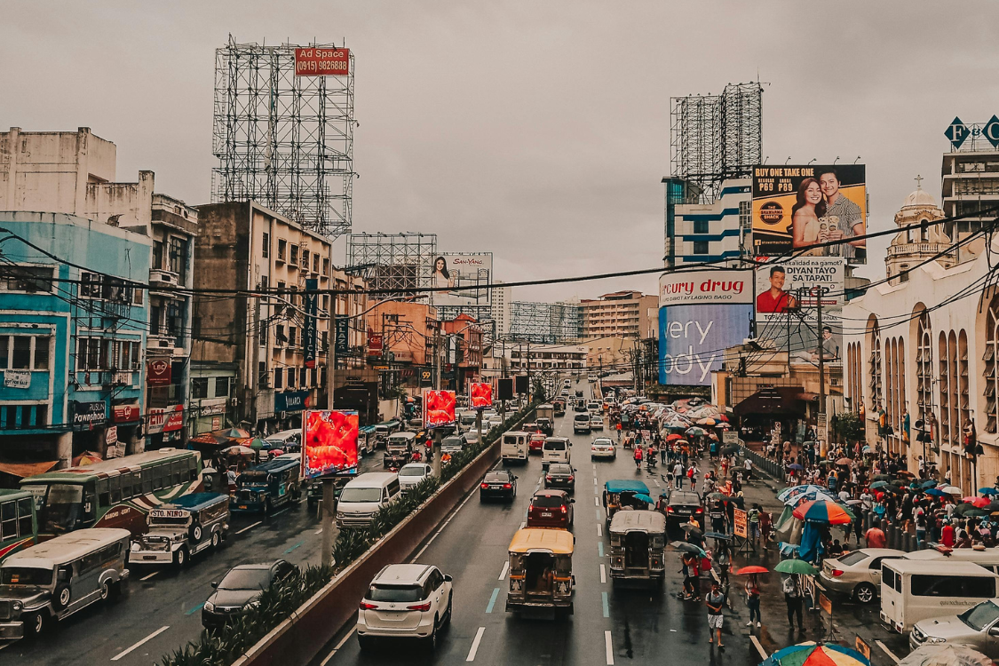 Busy street in Metro Manila with vehicles, pedestrians, and colorful signage under cloudy skies.