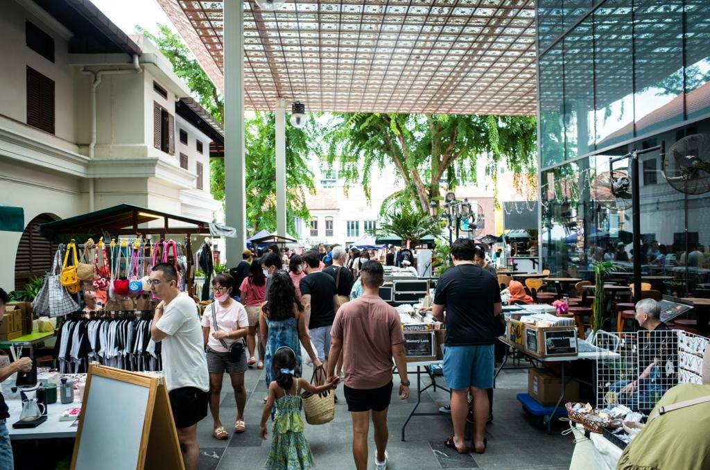 This image displays a lively outdoor market in Singapore sheltered by a large, modern canopy that filters sunlight onto the crowd. Shoppers browse through various stalls offering items like clothing, bags, and vinyl records while walking between historic white buildings and a sleek glass facade.
