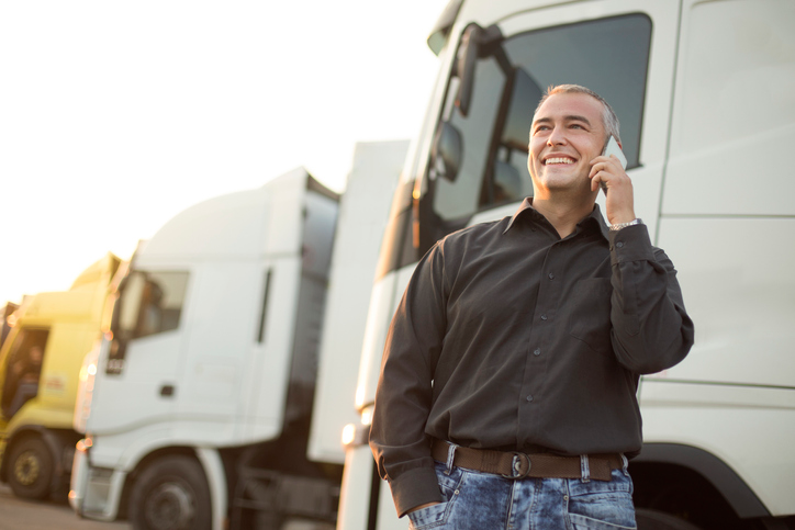 Man in jeans and a brown shirt talking on a cell phone next to a white truck. 