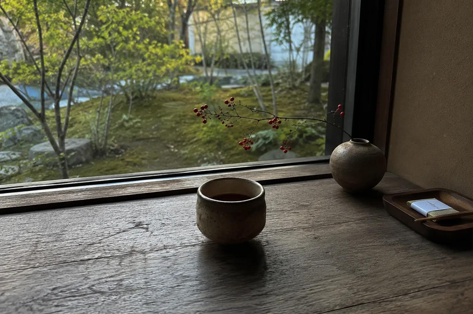 A close-up, slightly high-angle shot captures a serene tea setting on a rustic, textured wooden windowsill. In the foreground, a handmade, earth-toned ceramic cup filled with tea sits prominently, its surface showing natural variations in color. To the right, a round, textured clay vase holds a delicate, bare branch adorned with small red berries that stretches toward the left. Beside the vase is a small, dark wooden tray holding a packaged item with a toothpick. The large window in the background looks out onto a tranquil, moss-covered Japanese garden with green foliage and stone elements, softly blurred by a shallow depth of field.