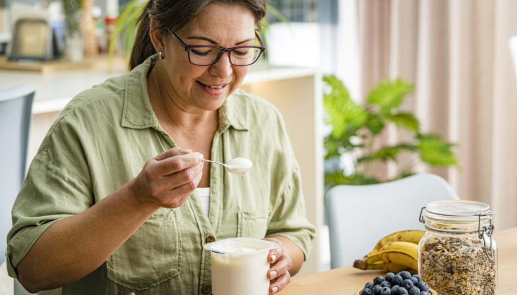 Kalsium – Derfor trenger kroppen din dette mineralet 14 Kvinne med briller smiler mens hun spiser yoghurt ved frokostbordet med bananer, blåbær og müsli i et lyst kjøkken.