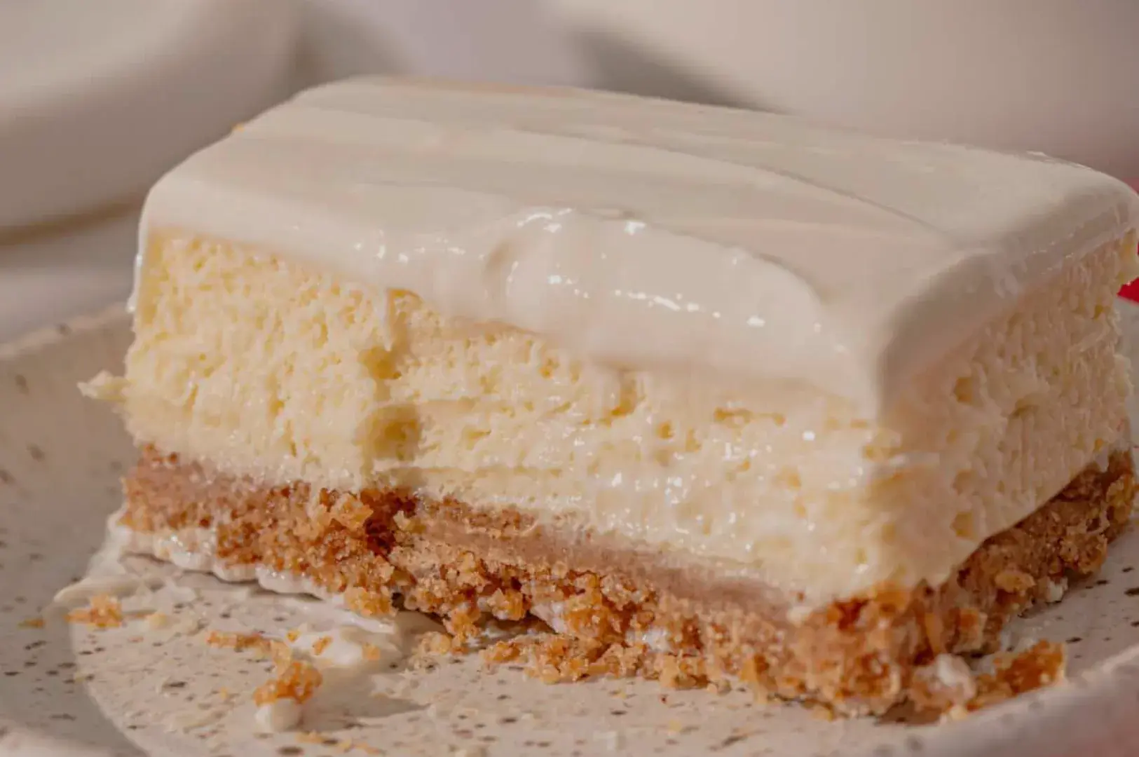 A slice of dessert with a creamy white topping, layered cheesecake, and a crumbly crust.
