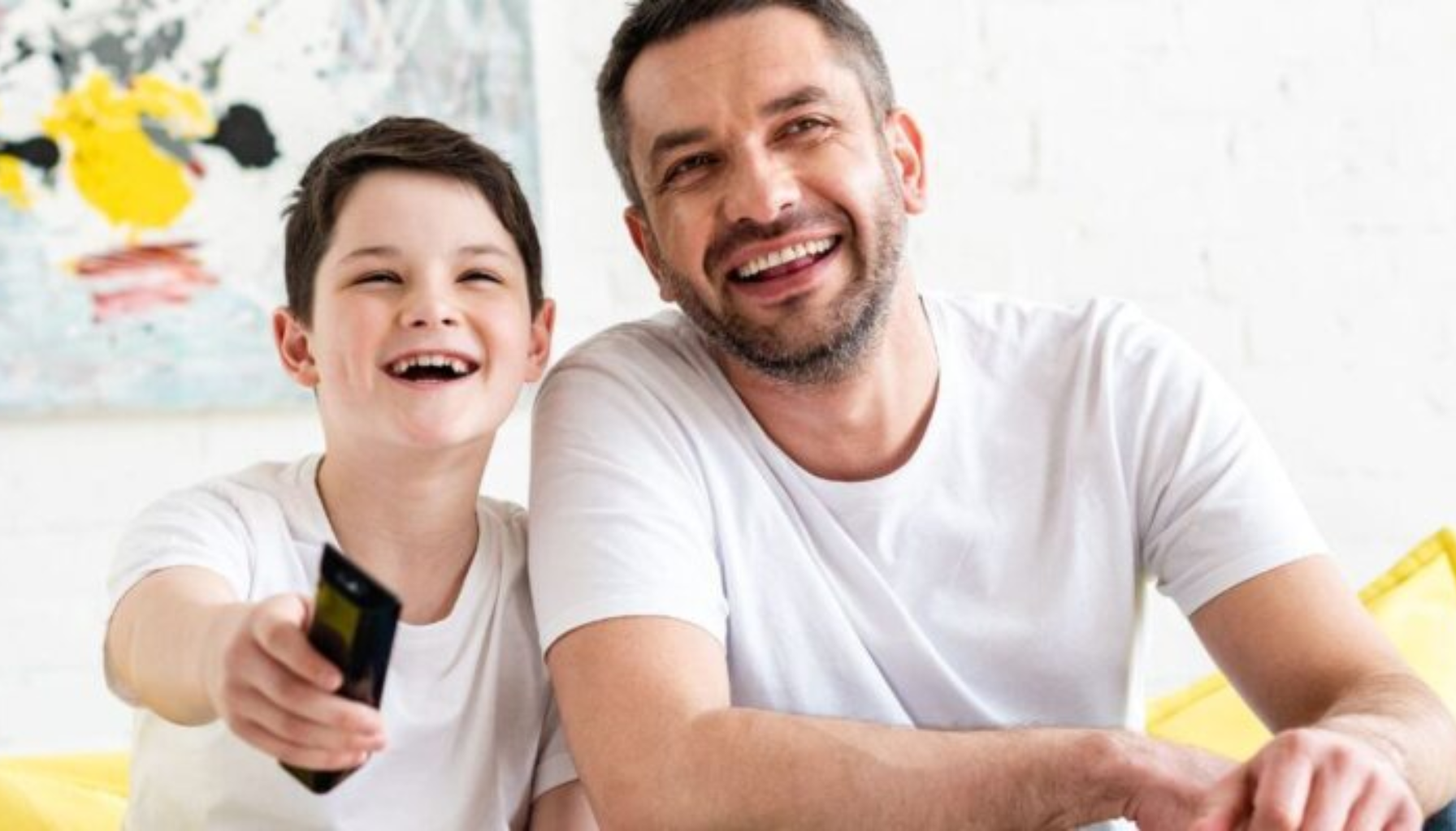 "Far og sønn sitter sammen i sofaen, smiler og ser på TV med fjernkontroll."