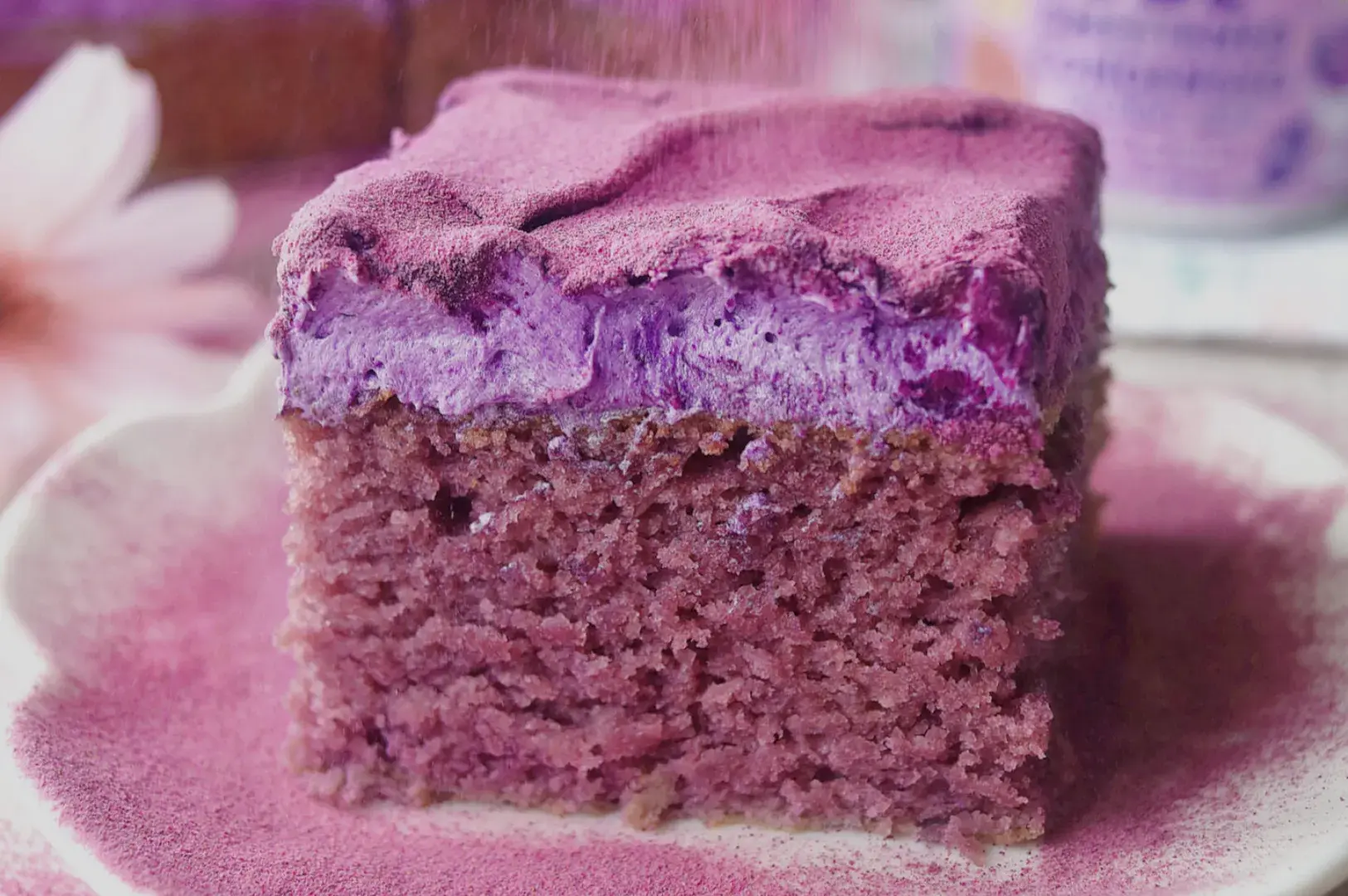 A slice of purple cake with a vibrant purple frosting, sitting on a plate with a sprinkle of purple dust.