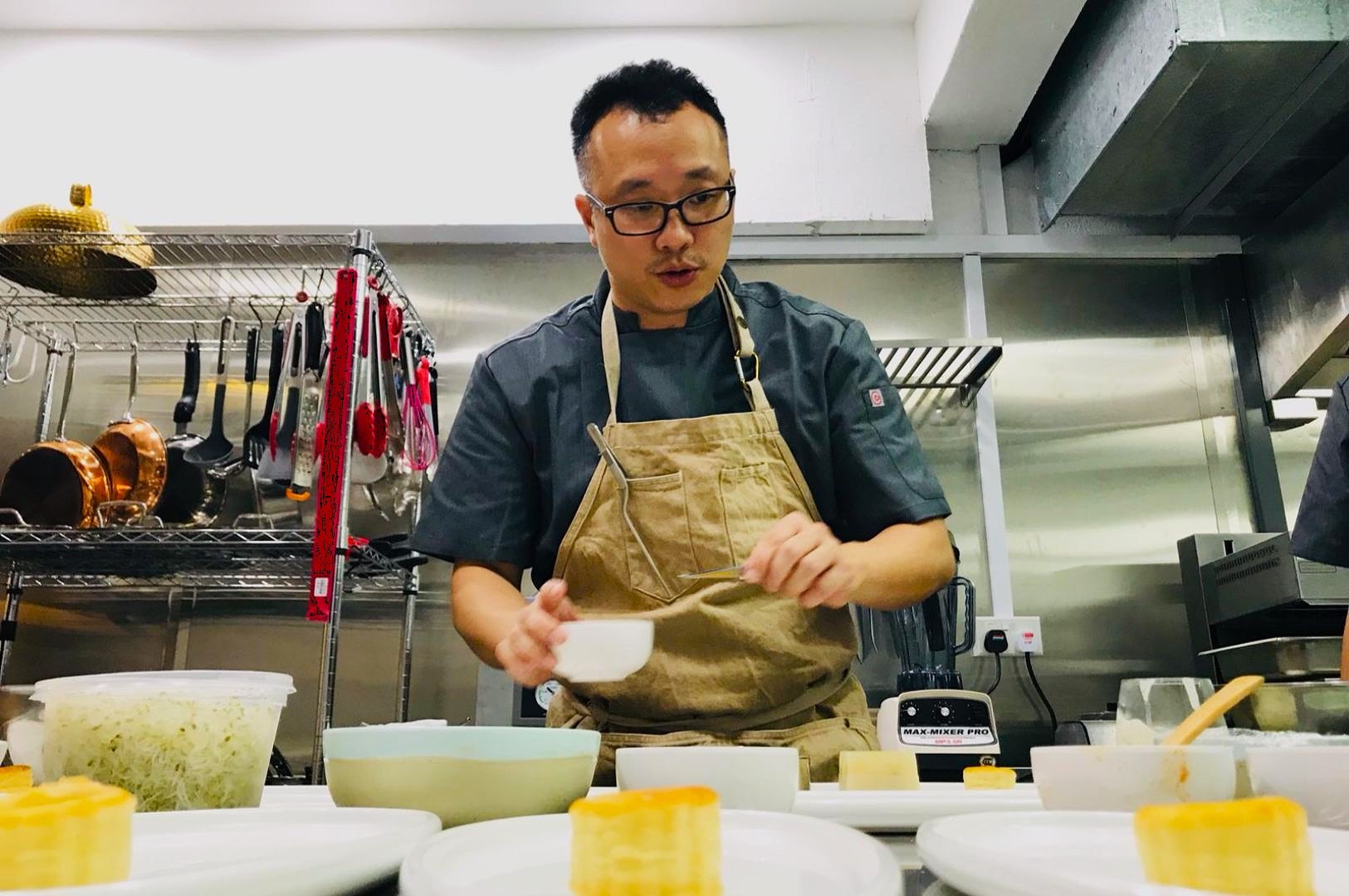 Chef wearing an apron, focused on preparing dishes in a professional kitchen. Shelves with utensils and cookware are visible in the background.