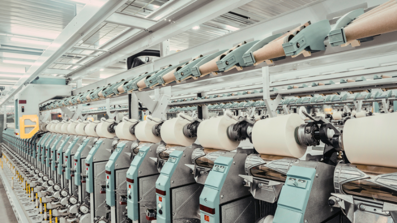 Rows of industrial spinning machines with large spools of white thread and cooling systems, viewed from an angle in a factory setting.