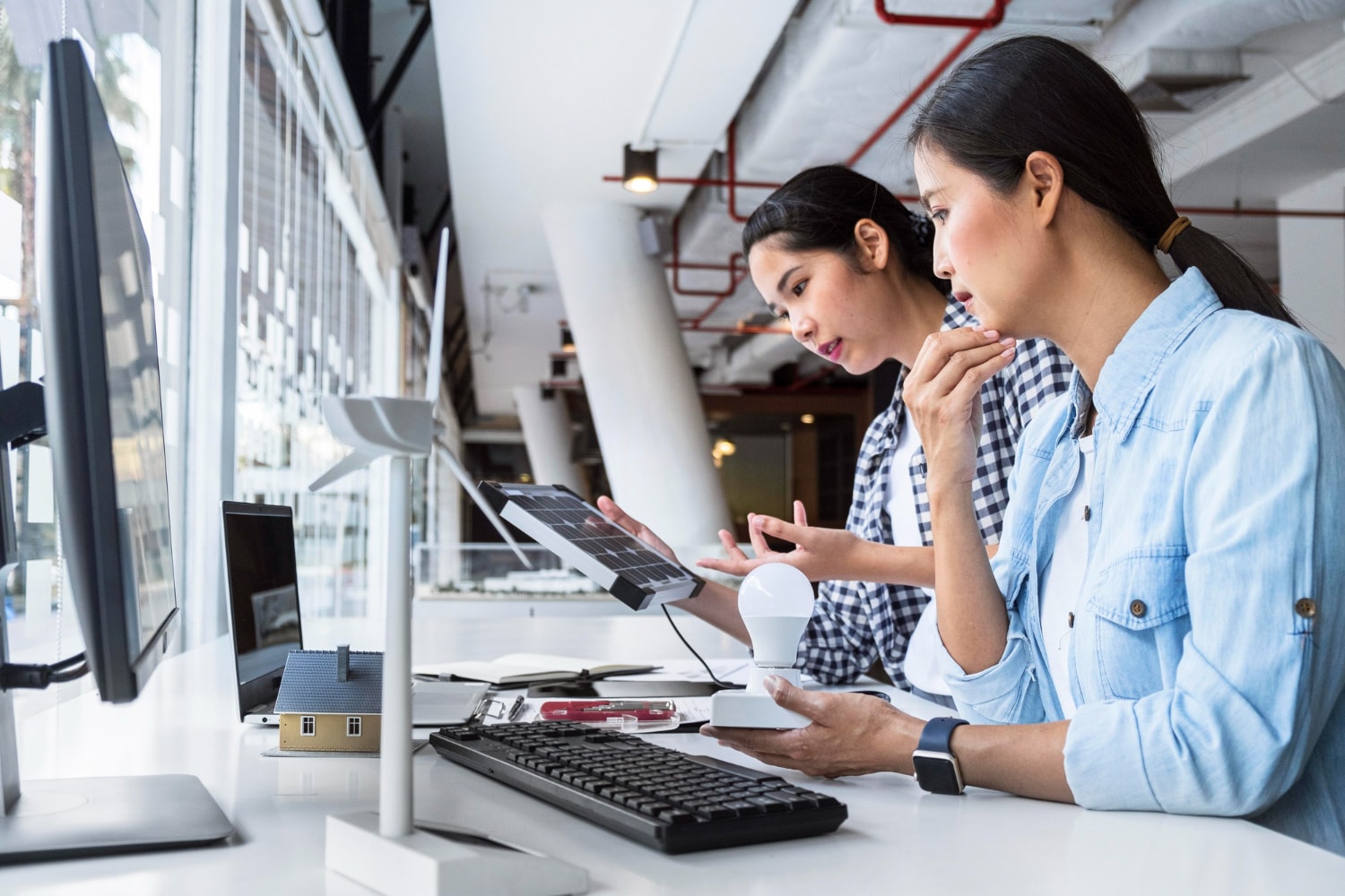 Two women test a solar panel and a smart lightbulb at a modern workstation, surrounded by desktop monitors and energy prototypes. UCaaS fosters innovation through unified communications tools that support cloud-based communication between R&D teams and remote collaborators via cloud telephony.