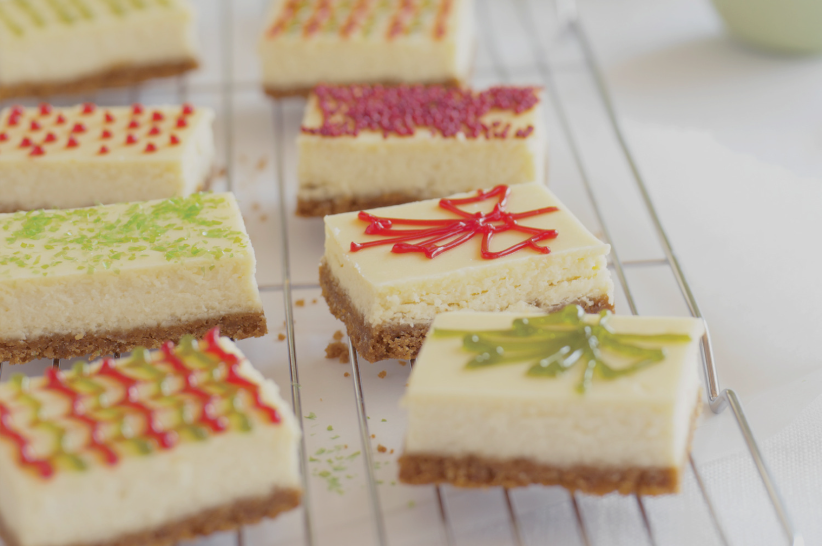 Assorted cheesecake bars on a cooling rack, topped with colorful decorations like green sprinkles and intricate red icing patterns, in a bright setting.