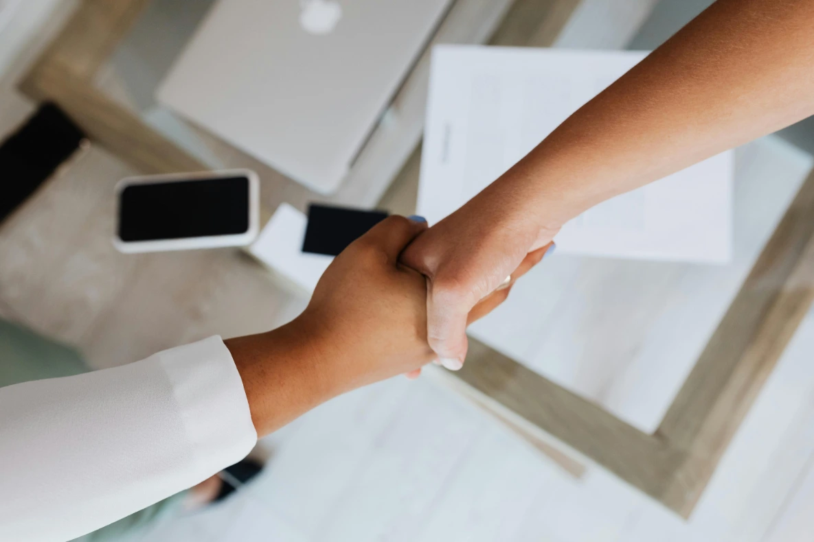 A handshake over a desk with a laptop, smartphone, and documents.