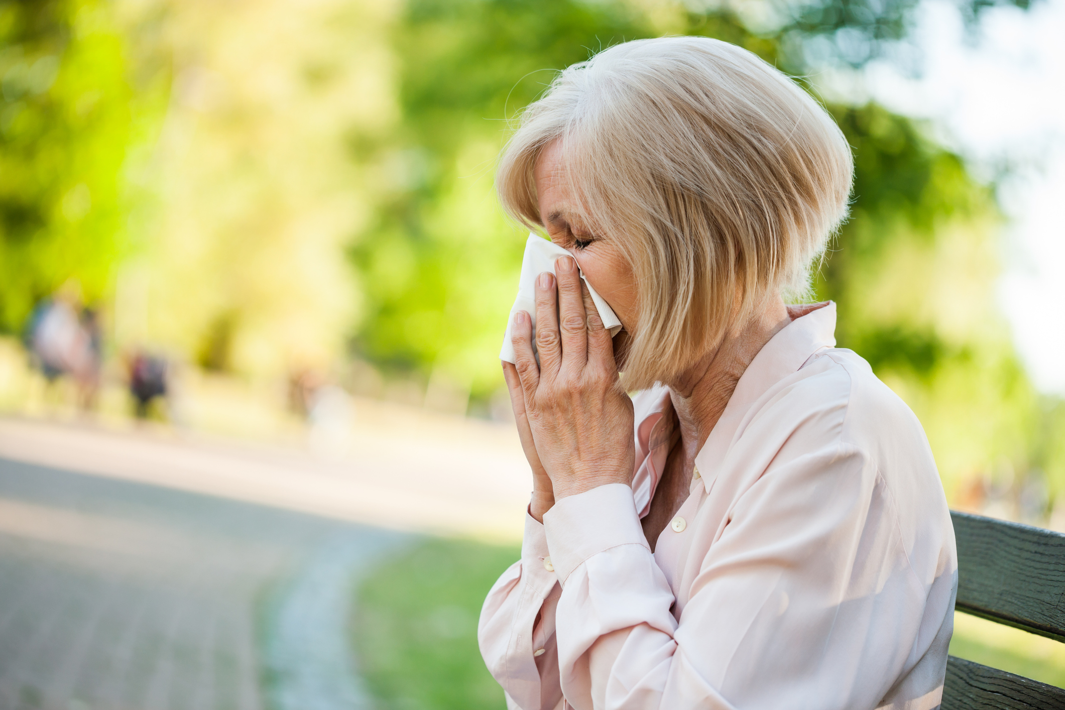 An image of a woman with allergies blowing her nose in a park. 